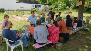 Vierzehn Menschen sitzen an einem langen Tisch im Garten. Sie essen und lachen.